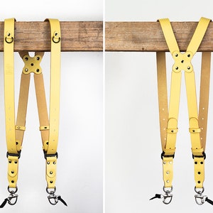 May include: Yellow leather camera strap with two adjustable straps and two metal hooks. The strap is hanging from a wooden surface.