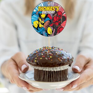 May include: A round cake topper with a picture of Wolverine and Deadpool. The topper says "Thomas's Birthday".