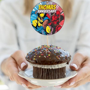 May include: A chocolate cupcake with colorful sprinkles topped with chocolate frosting. A round cake topper with a picture of Wolverine and Deadpool says "THOMAS ANNIVERSAIRE".