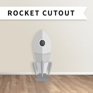 May include: A grey rocket cutout with a black window and a white background. The rocket is standing on a wooden floor.