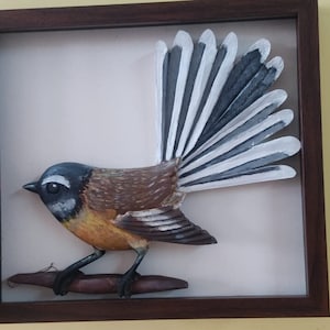 May include: A wooden carving of a bird with a long, fan-shaped tail. The bird is painted in shades of brown, gray, white, and yellow. The carving is displayed in a brown frame.