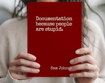 May include: A red book with the text "Documentation because people are stupid" and the author's name, Sam Johns, in white. The book is held by a person wearing a white jumper.