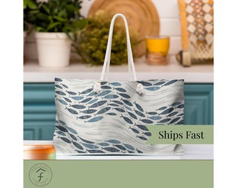 Könnte beinhalten: Eine große Tragetasche mit weißem Seilgriff. Die Tasche hat ein sich wiederholendes Muster aus blauen Fischen, die in einem wellenförmigen Design auf cremefarbenem Hintergrund schwimmen. Der Text "Ships Fast" ist auf der Tasche zu sehen.