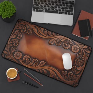 May include: A brown leather desk mat with an embossed floral design. The mat has a central panel and a decorative border with flowers and leaves. A white mouse sits on the mat, with a laptop and coffee cup nearby.