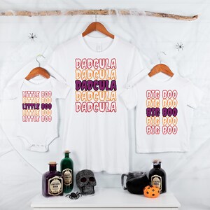 Puede incluir: Camisetas blancas y un body con texto tem&aacute;tico de Halloween. La camiseta de adulto dice "DADCULA" en letras apiladas de color naranja, morado y rosa. La camiseta infantil dice "BIG BOO", y el body dice "LITTLE BOO".