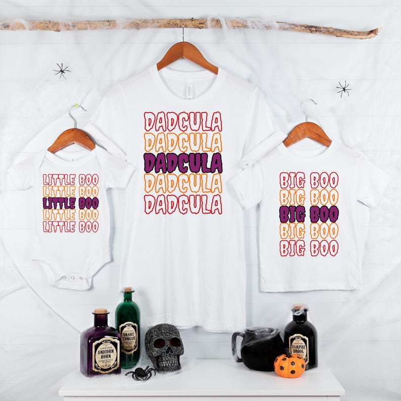 Puede incluir: Camisetas blancas y un body con texto tem&aacute;tico de Halloween. La camiseta de adulto dice "DADCULA" en letras apiladas de color naranja, morado y rosa. La camiseta infantil dice "BIG BOO", y el body dice "LITTLE BOO".