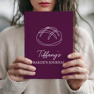 Op de afbeelding: Een dieppaars Baker's Journal met een witte lijntekening van een brood. De kaft van het tijdschrift heeft de tekst "Tiffany's BAKER'S JOURNAL" in witte letters. Het tijdschrift wordt door een persoon vastgehouden.