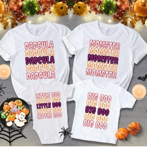 Puede incluir: Camisetas blancas con tem&aacute;tica de Halloween y un body con las frases "DADCULA", "MOMSTER", "LITTLE BOO" y "BIG BOO" en texto superpuesto naranja, morado y rojo. Las camisetas est&aacute;n dispuestas con decoraciones de Halloween.