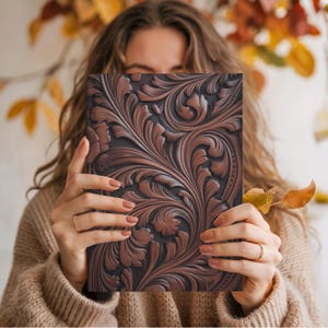 May include: A brown leather journal with an intricate floral pattern. The cover features a detailed embossed design with a dark brown background and lighter brown highlights. The journal is held in front of a person.