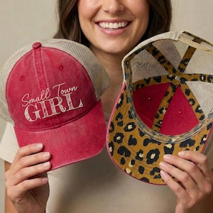 May include: Two red trucker hats with mesh backs. One hat features the text "Small Town GIRL" in white script. The other hat has a leopard print design on the underside of the brim. Both hats have a distressed, vintage look.