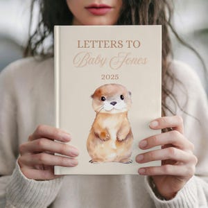May include: A cream-colored journal titled "Letters to Baby Jones 2025" with a watercolor illustration of a cute otter. The journal is held in front of a person wearing a cream-colored sweater.