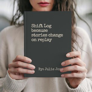 May include: A dark gray book titled "Shift Log because stories change on replay" by Julie Jor. The book is held by a person wearing a cream-colored sweater. The text is in a typewriter-style font.