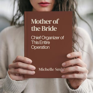 May include: A brown book titled "Mother of the Bride" with the subtitle "Chief Organizer of This Entire Operation." The author's name, Michelle Smith, is printed at the bottom. The book is held in front of a person.