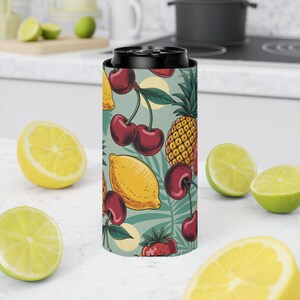 Puede incluir: Un enfriador de latas negro con un patrón colorido de cerezas, limones, piñas y fresas sobre un fondo turquesa. El diseño incluye hojas verdes y círculos amarillos. El enfriador está rodeado de limones y limas cortadas.