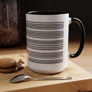 Könnte beinhalten: Weißer Keramikbecher mit schwarzem Innenraum, Henkel und einem sich wiederholenden schwarz-weißen geometrischen Muster. Der Becher steht auf einer Holzoberfläche mit einem Löffel und einem Macaron.