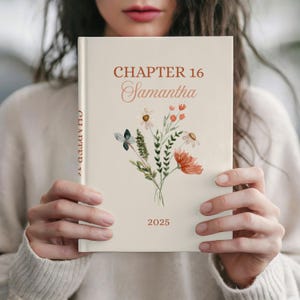 May include: A cream-colored book with the text "CHAPTER 16 Samantha 2025" in a reddish-brown font. The cover features a watercolor floral design with daisies, orange flowers, and greenery. The spine reads "CHAPTER 16".