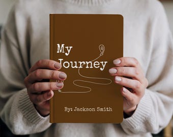 May include: A brown journal titled "My Journey" with white text and a line art design. The author's name, Jackson Smith, is printed at the bottom. The journal is held by a person wearing a cream-colored sweater.