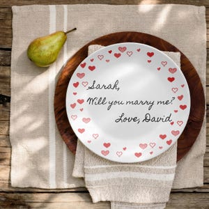 May include: A white plate with red heart designs and the words "Sarah, Will you marry me? Love, David" in black script. The plate sits on a wooden charger and a beige napkin. A green pear is on the left.