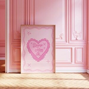 Pode incluir: Um pôster rosa e branco com um design em forma de coração e o texto "Dump Him Sis" em uma fonte rosa cursiva. O pôster é emoldurado em uma moldura dourada e está pendurado em uma parede rosa.