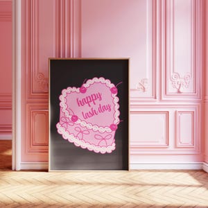 May include: A framed print featuring a pink heart-shaped cake design with the text "happy lash day" in a playful font. The cake is adorned with cherries. The print is set against a black background, framed in a wooden frame, and displayed in a pink room.