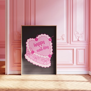以下が含まれることがあります： 「happy nail day」の文字が入った、ピンク色のハート型ケーキのデザインのフレームプリント。ケーキはさくらんぼとリボンで飾られ、黒い背景に配置されています。プリントは、ピンク色の壁と木製の床がある部屋に展示されています。