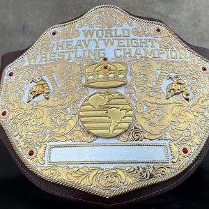 Puede incluir: Un cinturón de campeón mundial de lucha de peso pesado de oro y plata con respaldo de cuero marrón. El cinturón presenta adornos de volutas, una corona y un diseño de globo. Joyas rojas están engastadas en los bordes.