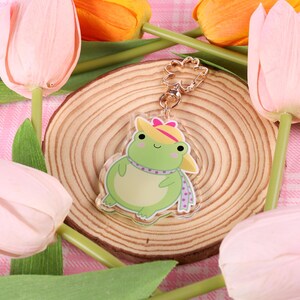 May include: A green frog wearing a pink and white hat and a purple scarf. The frog is a charm on a gold chain. The charm is sitting on a wooden surface with pink tulips in the background.