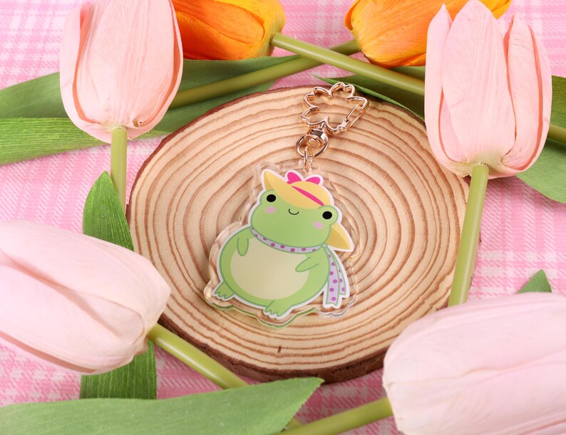 May include: A green frog wearing a pink and white hat and a purple scarf. The frog is a charm on a gold chain. The charm is sitting on a wooden surface with pink tulips in the background.