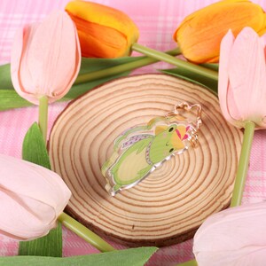 May include: A green frog charm with a pink crown, on a wooden coaster, surrounded by pink and orange tulips.