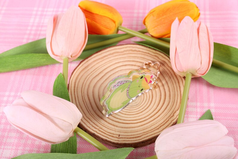 May include: A green frog charm with a pink crown, on a wooden coaster, surrounded by pink and orange tulips.