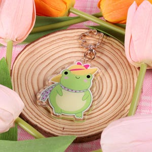 May include: A green frog wearing a yellow hat and a purple scarf. The frog is a keychain charm with a gold metal ring.