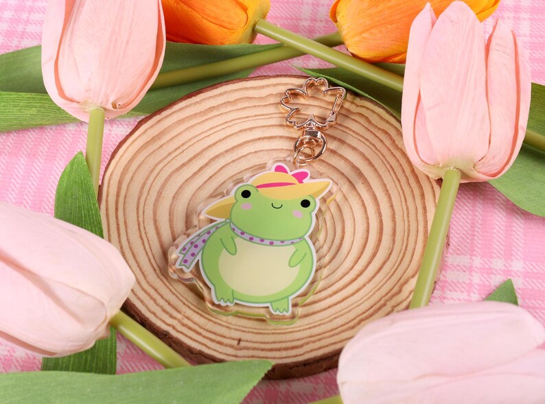 May include: A green frog wearing a yellow hat and a purple scarf. The frog is a keychain charm with a gold metal ring.