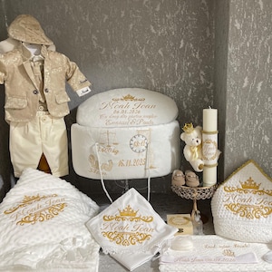 Pode incluir: Um conjunto de artigos de batismo para bebé em creme e dourado. Inclui um fato, um cobertor, uma toalha, um chapéu e uma vela, todos bordados com o nome "Noah Joan" e um desenho de coroa. O conjunto também apresenta um ursinho de peluche e sapatos.