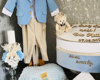 Op de afbeelding: Een formele outfit voor een babyjongen in lichtblauw, beige en wit, inclusief een blazer, broek, overhemd, vlinderdas en hoed. Een teddybeer en een taart met tekst zijn ook aanwezig, samen met een deken en kussen.