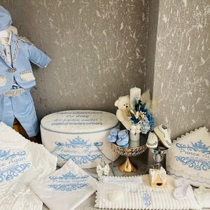 Pode incluir: Um conjunto de batismo para bebé em azul claro e branco. O conjunto inclui um fato, chapéu, cobertor, almofada e outros acessórios, todos decorados com uma coroa e o nome "Prince Ayan".