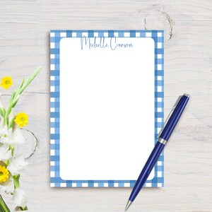May include: Blue and white gingham notepad with a white border and the name "Michelle Carson" printed at the top. A blue pen is lying on the notepad.
