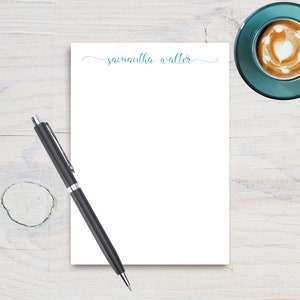 May include: A white notepad with the text "samantha walter" in blue cursive script. A black pen is lying on the notepad. A cup of coffee with a heart design in the foam is in the top right corner of the image.
