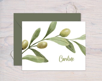May include: A white card with a watercolor illustration of an olive branch with green leaves and olives. The name "Caroline" is written in cursive at the bottom of the card.