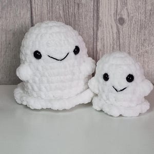 Könnte beinhalten: Zwei weiße Häkelgespenster mit schwarzen Knopfaugen und lächelnden Mündern. Das größere Gespenst hat einen abgerundeten Körper und kleine Arme. Das kleinere Gespenst hat ein ähnliches Design. Die Geister werden auf einer weißen Oberfläche mit Holzmaserung im Hintergrund gezeigt.