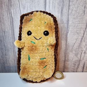 May include: A brown and tan plush toy shaped like a piece of toast with a smiling face and green embroidery. The toy is about the size of a UK one pound coin.