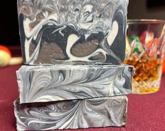 May include: Three bars of handmade soap stacked on top of each other. The soap is a swirl of black, white, and gray. The soap is sitting on a red surface.