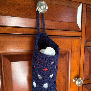 May include: A blue crocheted bag with white and red accents hangs from a cabinet door knob. The bag is filled with white plastic bags.