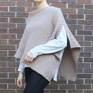 May include: A brown knitted poncho with a loose fit and a split in the back. The poncho is worn over a white long-sleeved shirt and black jeans.