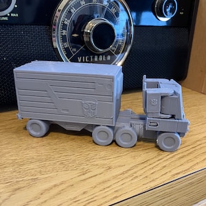 Puede incluir: Un camión de juguete gris con un remolque. El camión tiene una cabina grande y un remolque de plataforma. El camión está hecho de plástico y tiene un diseño detallado. El camión está sentado sobre una superficie de madera.