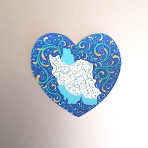 May include: A heart-shaped sticker with a blue background and a decorative pattern of swirls and leaves in green and gold. The sticker features a light blue outline of Iran, filled with a white and gold swirl design.