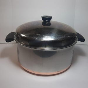 May include: A stainless steel stockpot with a copper-colored base and a matching lid. The pot features two black side handles and a black knob on the center of the lid. The exterior surface is reflective, showing a metallic silver finish.