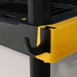 May include: Close-up of a black and yellow shelving unit corner. The corner features a black hook-shaped component that connects to a black support pole. The shelf edge is yellow, and the shelf itself is black.