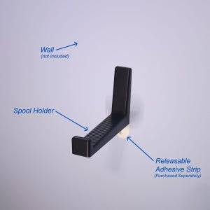 May include: A black spool holder with a ridged surface, mounted on a white wall. The holder is attached with a releasable adhesive strip. The text on the image reads "Spool Holder" and "Releasable Adhesive Strip (Purchased Seperately)".