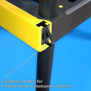 May include: Close-up of a black carabiner holder attached to a yellow and black shelf corner. The text "Carabiner Holder for Project Source Commander Shelves" is visible at the bottom of the image. The shelf has a black leg.