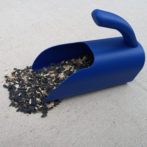 May include: A blue plastic scoop with a handle, filled with birdseed, spilling out onto a light gray surface.